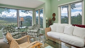 A living room with a glass coffee table, two patterned armchairs, a white sofa, and a view of snow-capped mountains through large windows. - Deering Crest – 3BR Home with Panoramic RMNP Views – Windcliff – 20-NCD3249 (Estes Park)