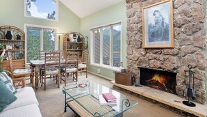 A cozy living room with a stone fireplace, wooden furniture, and large windows offering a view of the outdoors. - Deering Crest – 3BR Home with Panoramic RMNP Views – Windcliff – 20-NCD3249 (Estes Park)