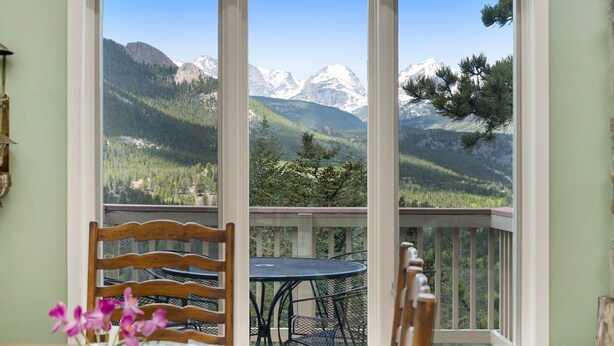 A balcony with a table and chairs, overlooking a mountainous landscape with snow-capped peaks and dense forest. - Deering Crest – 3BR Home with Panoramic RMNP Views – Windcliff – 20-NCD3249 (Estes Park)