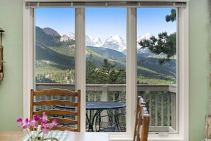 Uma varanda com mesa e cadeiras, com vista para uma paisagem montanhosa com picos nevados e floresta densa.