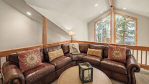 Una sala de estar acogedora con un sofá seccional de cuero marrón, una mesa redonda de centro y una ventana con vista a árboles.