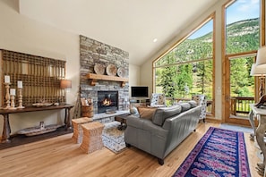 Un salon avec une cheminée en pierre, un canapé gris, une grande fenêtre donnant sur la forêt et un plancher en bois.