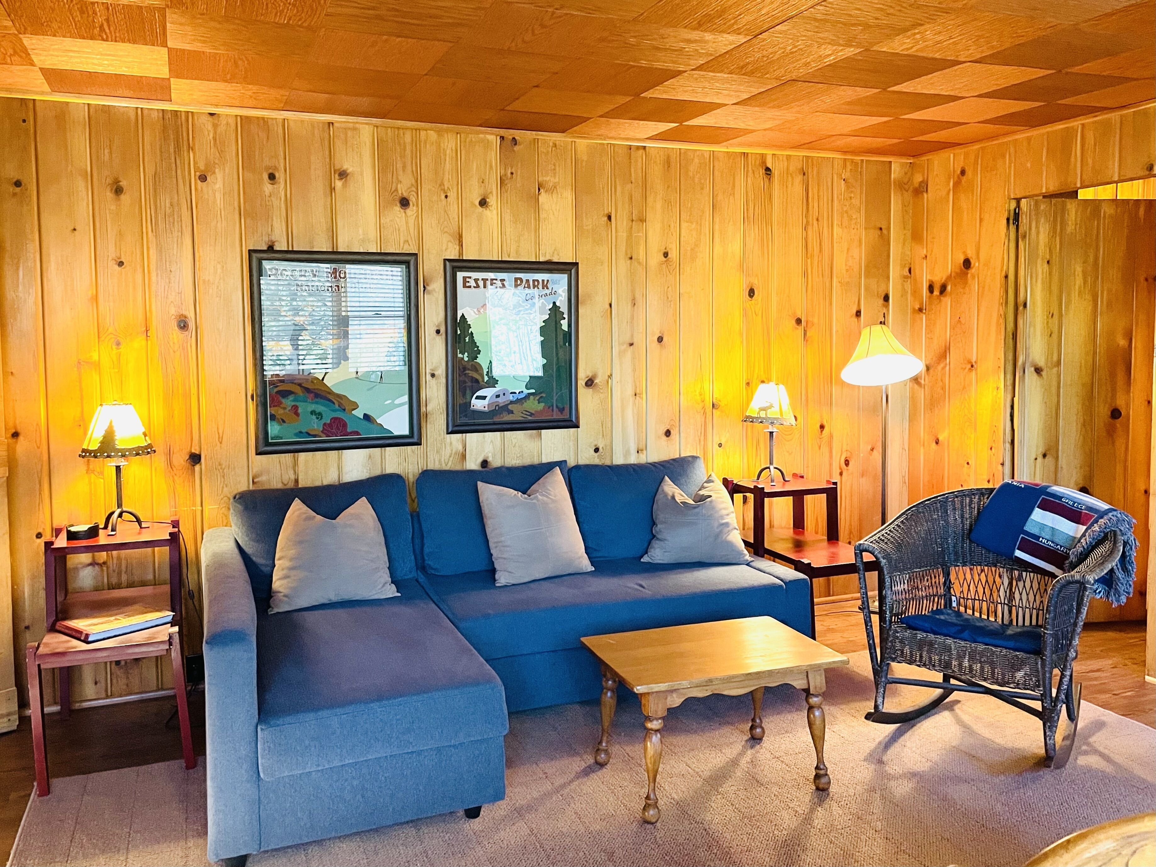 A cozy room with a blue sectional sofa, a wooden coffee table, a wicker rocking chair, and two framed posters on the wall.