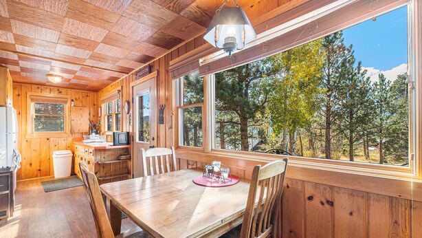 A wooden cabin interior with a dining table, chairs, a kitchen area, and large windows overlooking a forest.