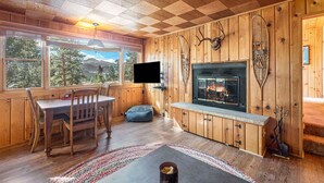 A cozy cabin interior with a wooden ceiling, a fireplace, a television, and a dining area with a table and chairs.