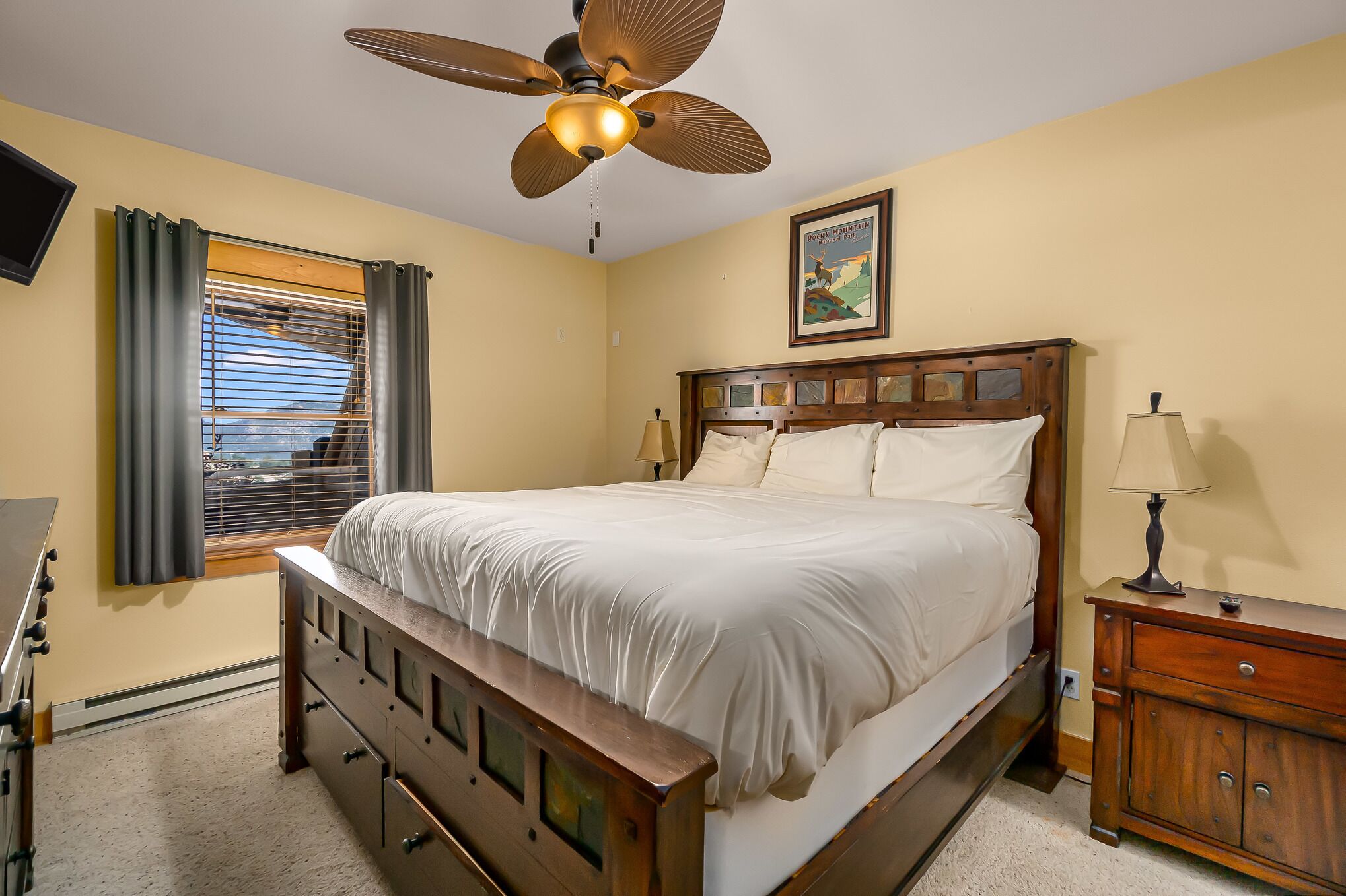 A bedroom with a bed, a wooden nightstand, a ceiling fan, a window with blinds, and a wall-mounted TV.