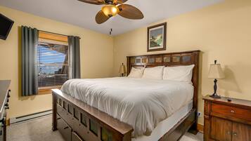 A bedroom with a bed, a wooden nightstand, a ceiling fan, a window with blinds, and a wall-mounted TV.