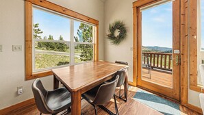 A dining area with a wooden table and chairs, large windows, a door leading to an outdoor deck, and a wreath on the wall.