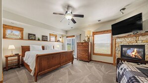 A bedroom with a wooden bed, a ceiling fan, a fireplace, a TV mounted on the wall, and a dresser.