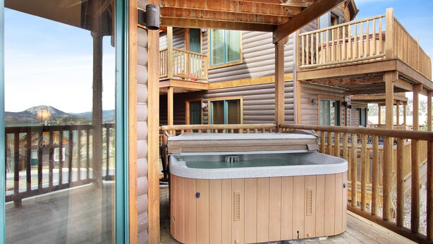 A wooden cabin with a hot tub on the deck, a balcony, and a mountain view.