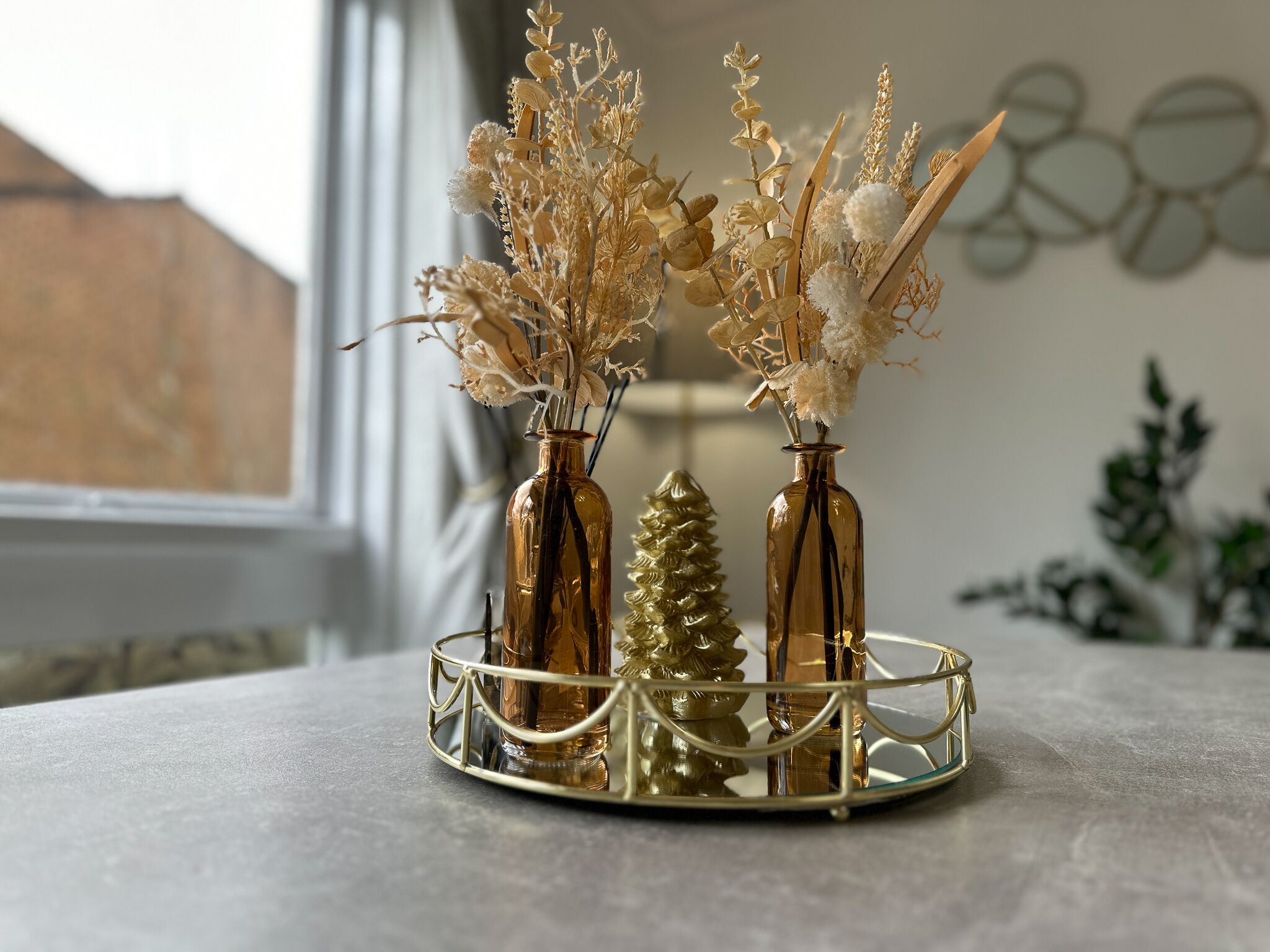Two glass vases with dried flowers on a round, gold-framed tray.