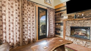 A cozy living room with a fireplace, a wooden table, and curtains with a leaf pattern. - Show Me Mountain Views – Gorgeous Home Near RMNP – 20-NCD0130 (Estes Park)