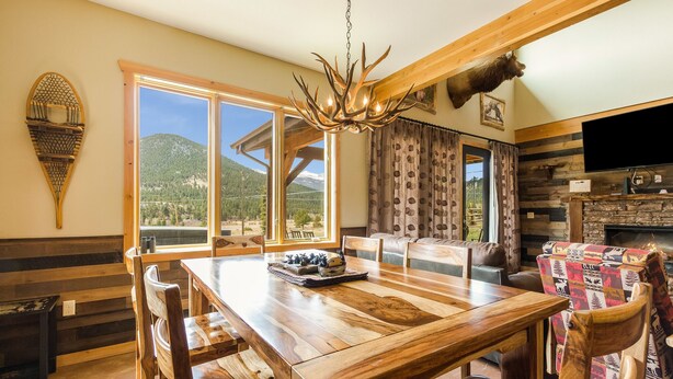 A wooden dining table with chairs, a chandelier, a mounted animal head, and a TV. - Show Me Mountain Views – Gorgeous Home Near RMNP – 20-NCD0130 (Estes Park)