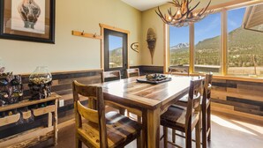 A dining area with a wooden table and chairs, a chandelier, and a mountain view through large windows. - Show Me Mountain Views – Gorgeous Home Near RMNP – 20-NCD0130 (Estes Park)