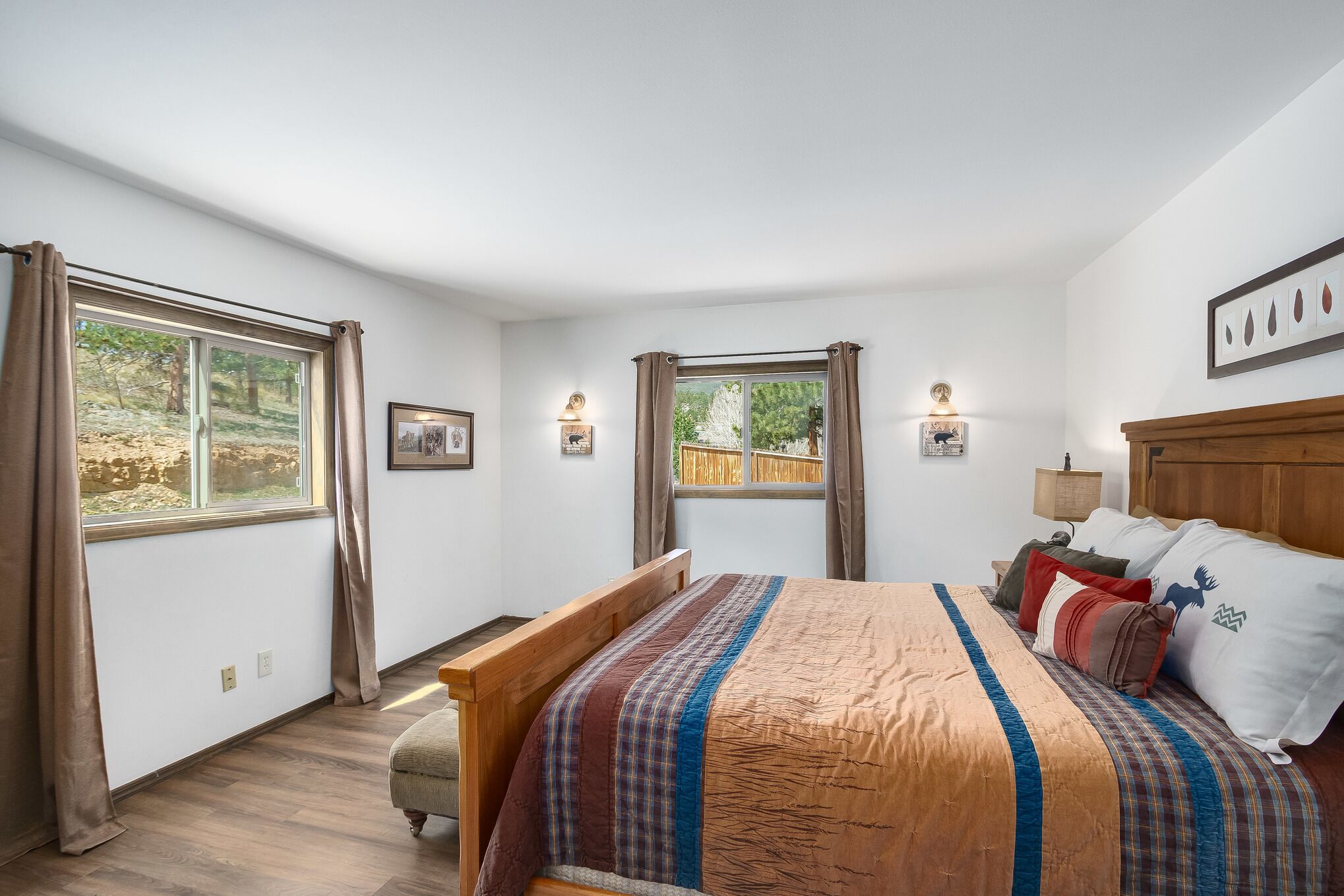 Un dormitorio con cama de madera, dos ventanas con cortinas, una mesita de noche con lámpara y un cuadro enmarcado en la pared.