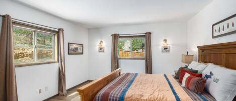 Un dormitorio con cama de madera, dos ventanas con cortinas, una mesita de noche con lámpara y un cuadro enmarcado en la pared.