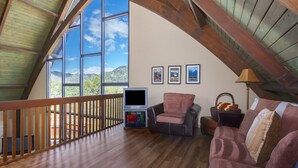 A spacious loft with a wooden floor, a large window offering a mountain view, a television, and a cozy seating area with a couch and armchair.