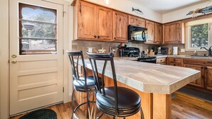 Une cuisine avec des meubles en bois, un grand îlot central doté de tabourets, un four à micro-ondes et une fenêtre donnant sur les arbres.