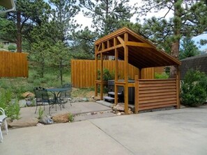 A wooden outdoor structure with a roof, a barbecue grill, and a table with chairs.