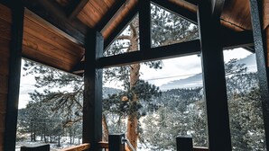 A wooden deck with a railing, a large window, and a view of snow-covered trees and mountains.