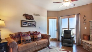 A living room with a brown sofa, patterned cushions, a ceiling fan, a fireplace, and a large window with curtains.