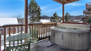 A wooden deck with a hot tub, a glass table, and a chair, overlooking a snowy landscape with mountains.