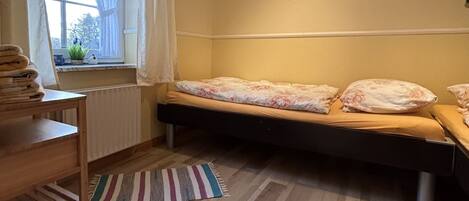 A small, cozy room with a bed, a radiator, a window with curtains, a small table with towels, and a colorful rug.