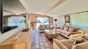 Un salon spacieux doté d’un grand téléviseur à écran plat, de sièges confortables, d’une table basse et offrant une vue sur l’extérieur grâce à de grandes fenêtres.