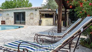 Un bâtiment en pierre doté d’une piscine, de chaises longues et d’une pergola en bois ornée de fleurs rouges.