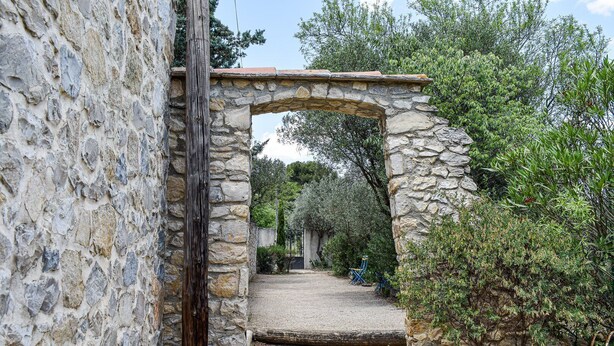 Un arc en pierre mène à une cour intérieure, où un chemin de gravier serpente au milieu de la verdure, le tout étant bordé d’un mur de pierre.