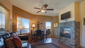 A living room with a fireplace, a ceiling fan, a television mounted above it, and a dining area with a table and chairs. - Mount Meeker 26B - Cozy Condo with Private Hot Tub - Mountain and Lake Views (Estes Park)