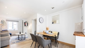 Un salon moderne comprenant un canapé, une table à manger avec des chaises, un téléviseur fixé au mur et une horloge.