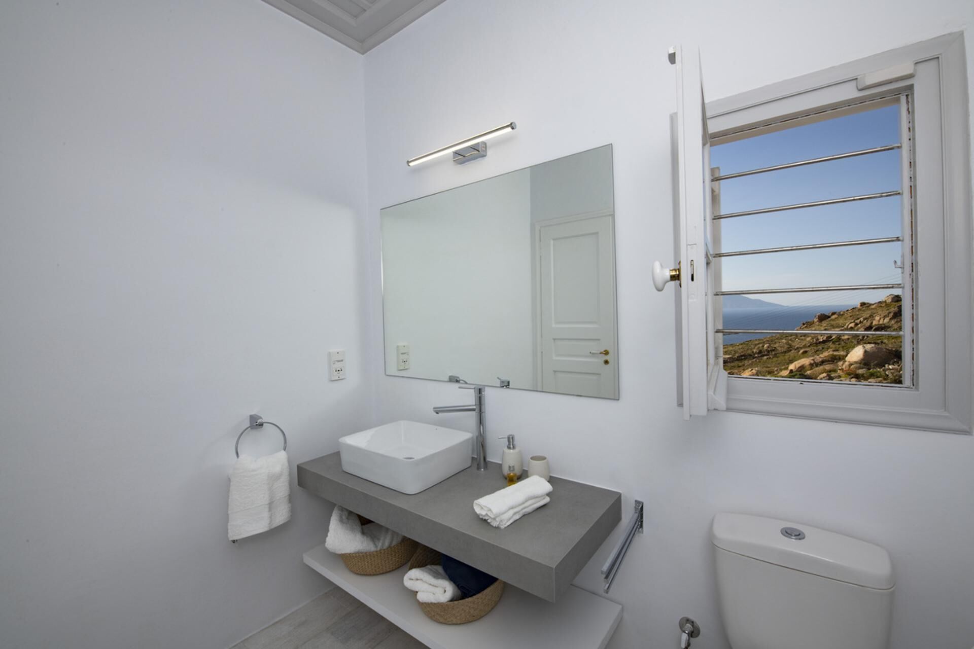 A modern bathroom with a white sink, a large mirror, and a window with a view of rocky terrain.