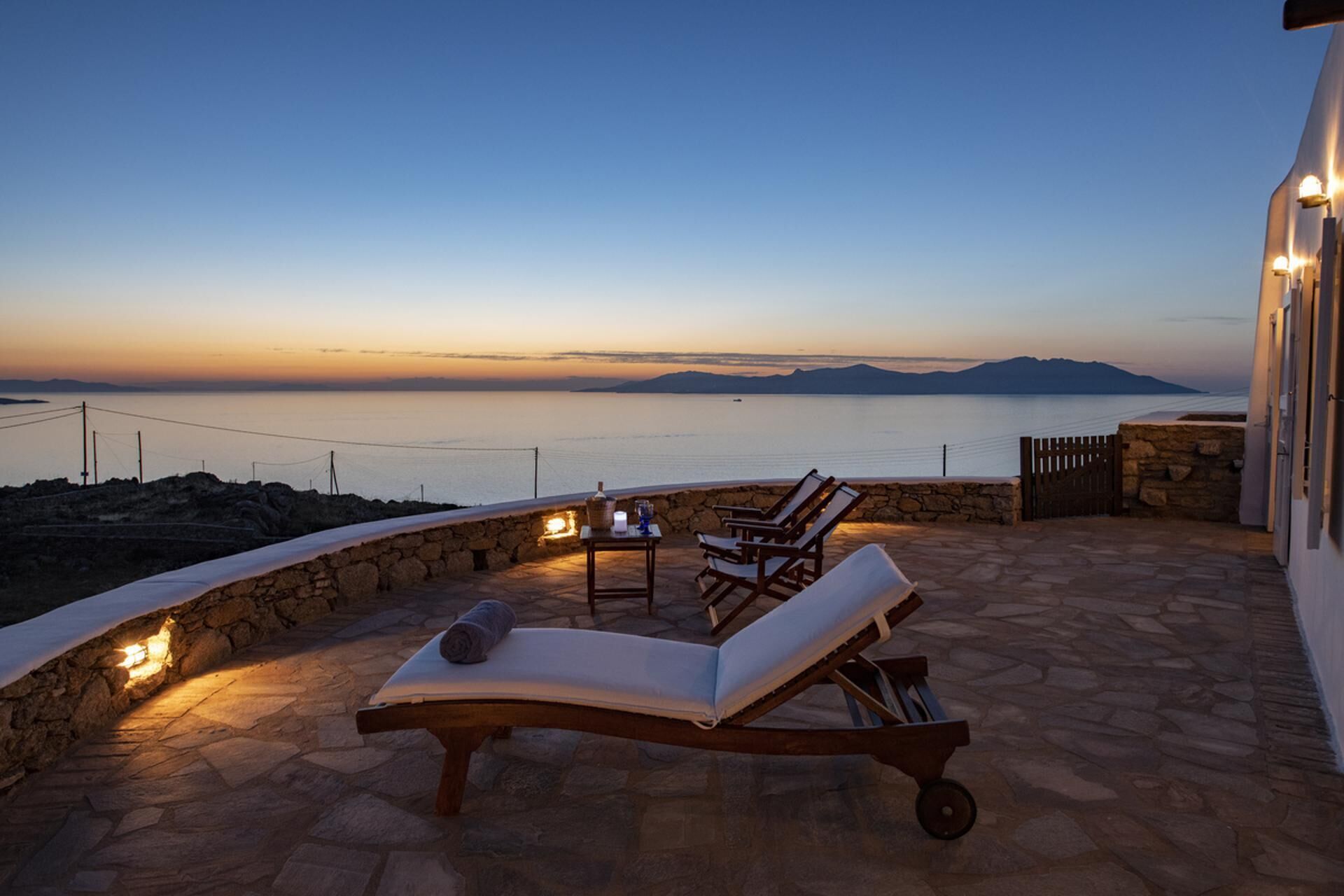 Une vue sur le coucher de soleil depuis une terrasse en pierre, avec des chaises longues et une table, donnant sur une mer calme et des montagnes lointaines.