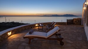 A sunset view from a stone patio with lounge chairs and a table, overlooking a calm sea and distant mountains. - Charming 5-bed Villa in Mykonos (Mykonos)
