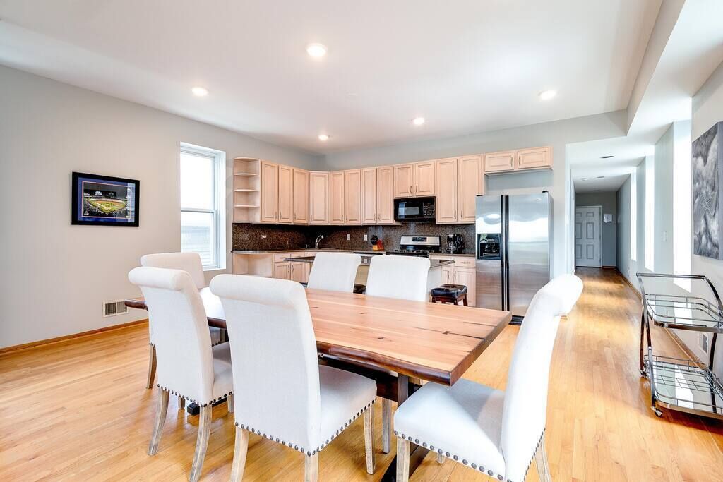 A modern kitchen with light wood cabinets, stainless steel appliances, and a dining area with white chairs and a wooden table.