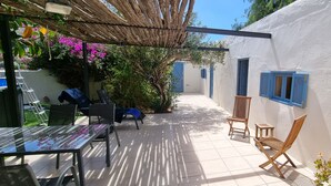 Un patio con una mesa y sillas, una pérgola y una pared blanca con contraventanas azules.