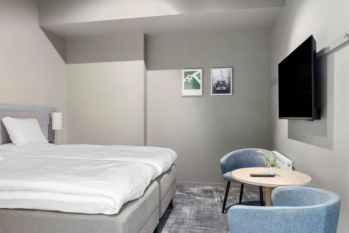 A hotel room with a bed, a small round table with a chair, and two framed pictures on the wall.