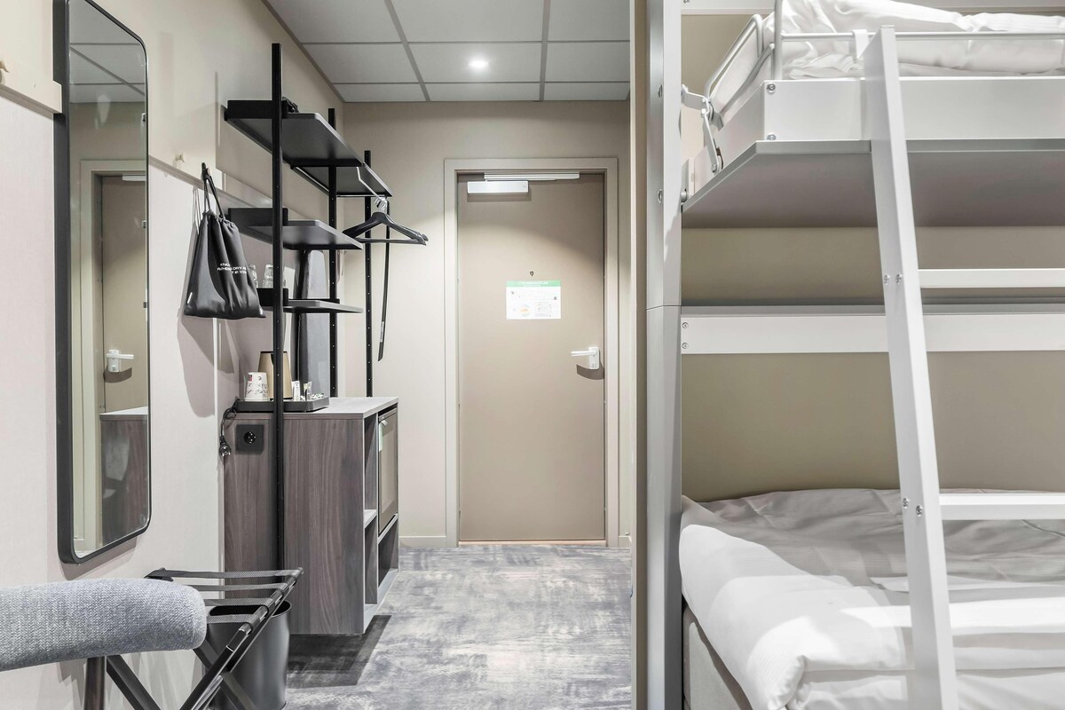 A hotel room with bunk beds, a desk with a chair, a mirror, a wardrobe, and a trash bin.