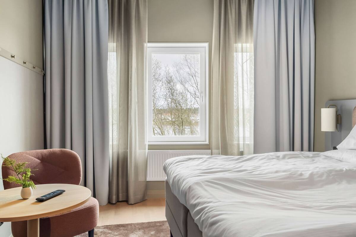 A hotel room with a bed, a chair, a small table with a plant and a remote control, and a window with curtains.