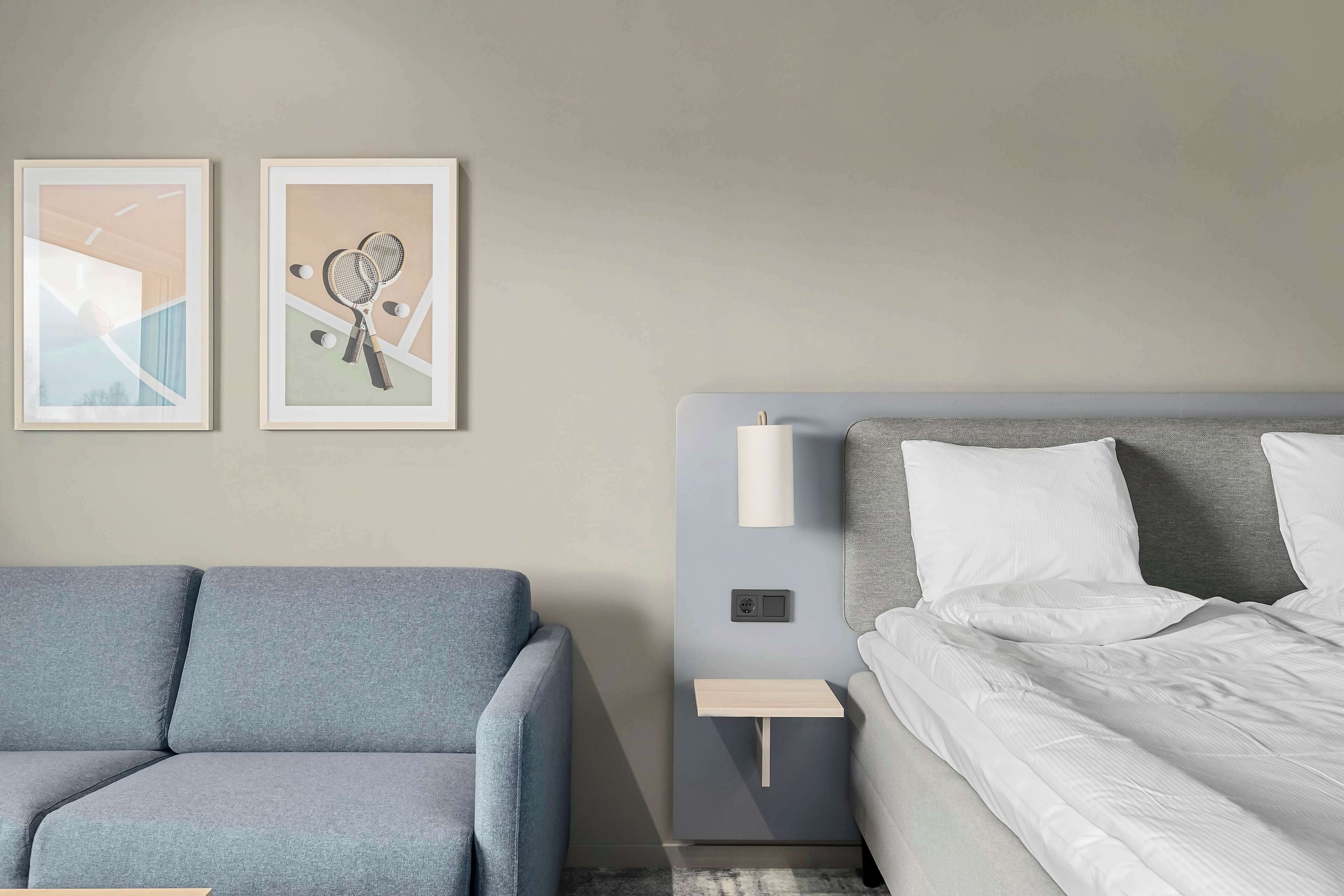 Ein modernes Hotelzimmer mit einem grauen Sofa, einem Bett mit weißer Bettwäsche und zwei gerahmten Bildern an der Wand.