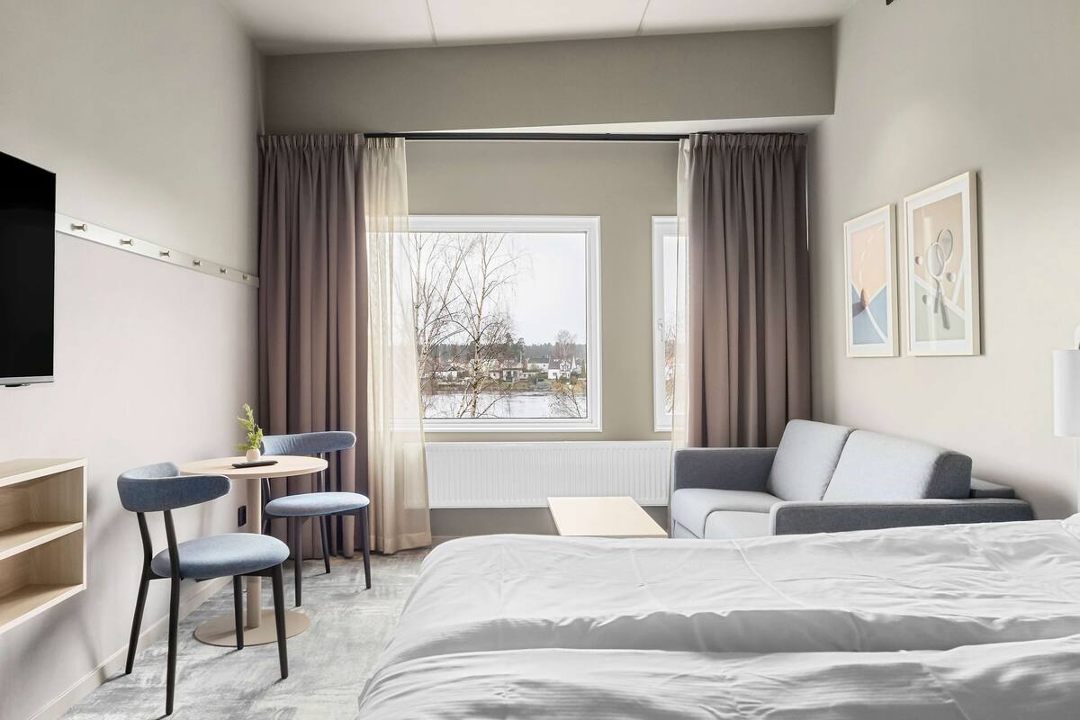 A hotel room with a bed, a sofa, a desk with chairs, a TV, and a window with curtains.