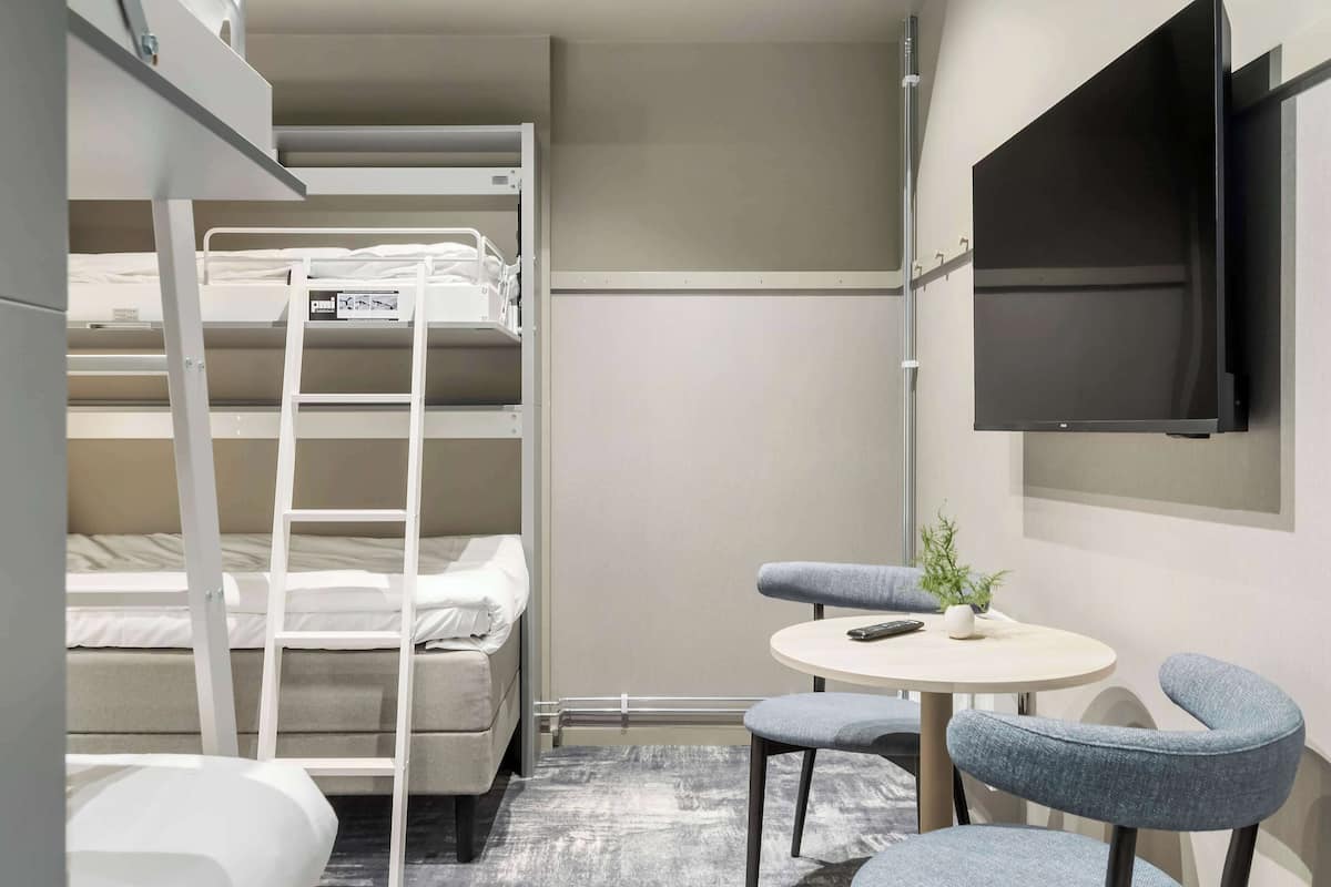 A compact hotel room with bunk beds, a small round table with a chair, a TV mounted on the wall, and a potted plant on the table.