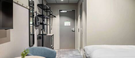 Habitación de hotel con una cama, una mesita redonda con una planta, una silla, un televisor montado en la pared y un armario con espejo.