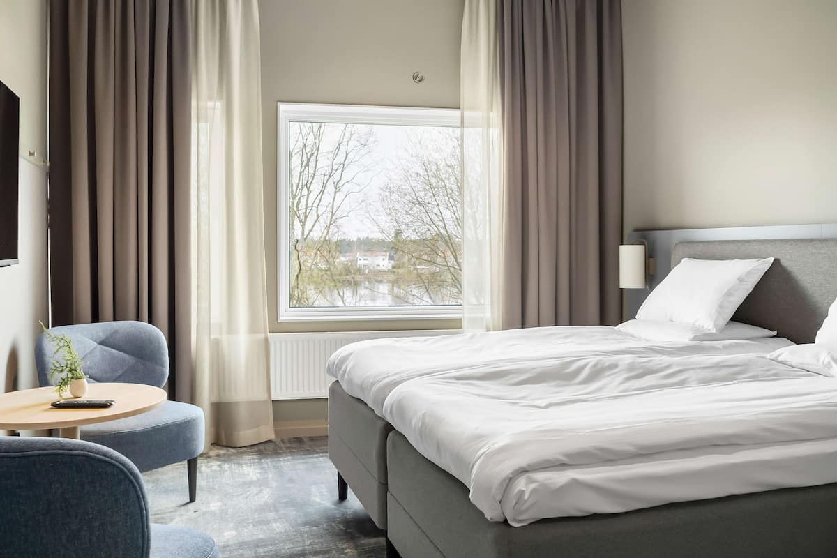 A hotel room with a bed, a round table with a remote control, a chair, and a window with curtains.