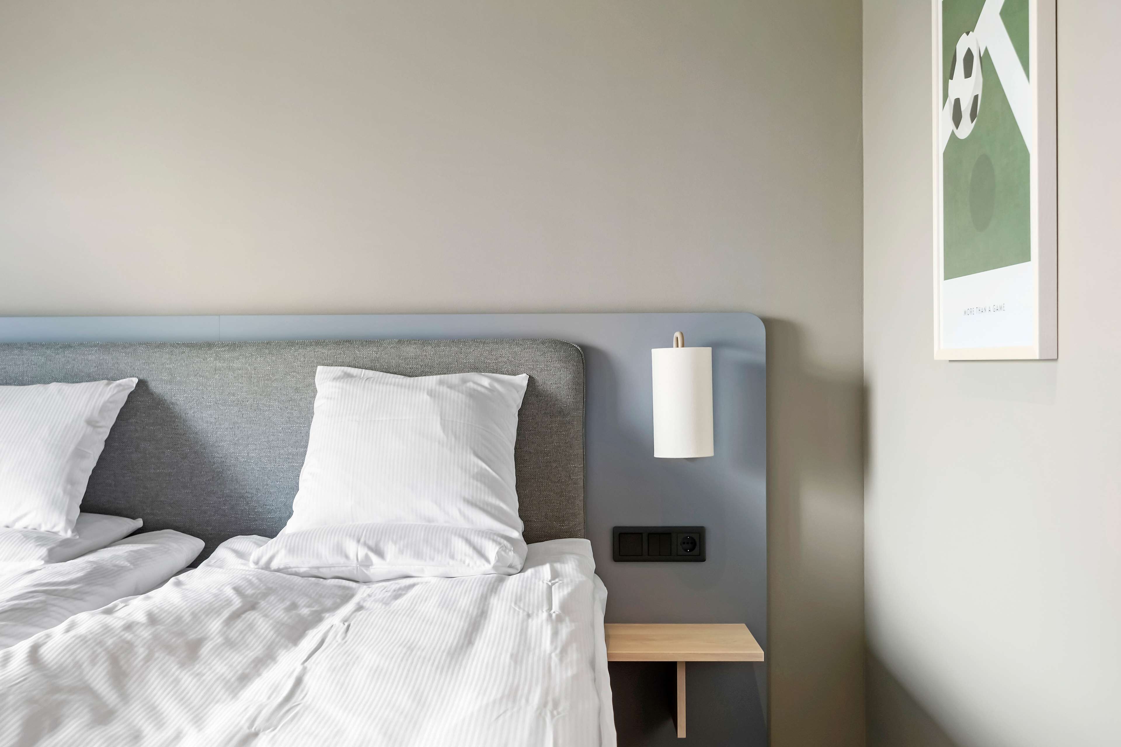 Una cama bien hecha con sábanas blancas, cabecera gris y mesita de noche con lámpara.