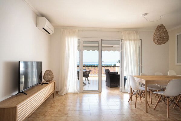 A modern living room with a TV, a wooden bench, a dining table with chairs, and a view of the sea through a glass door.