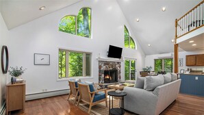A spacious living room with a fireplace, wooden flooring, and large windows offering a view of the outdoors. - Cozy Cottage w/ Shared Neighborhood Lake Access (Traverse City)