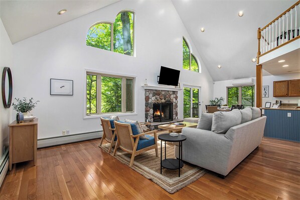 A spacious living room with a fireplace, wooden flooring, and large windows offering a view of the outdoors.