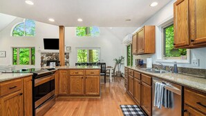 A spacious kitchen with wooden cabinets, a stone fireplace, and large windows offering views of greenery. - Cozy Cottage w/ Shared Neighborhood Lake Access (Traverse City)